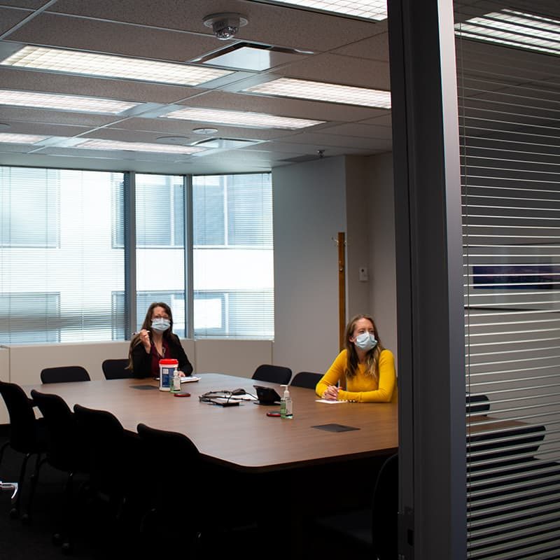 Two women, sitting in a boardroom and wearing blue masks