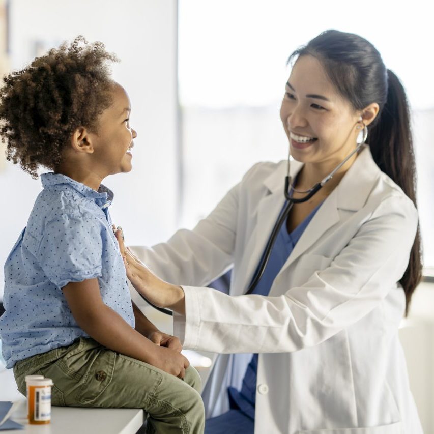 A female doctor of Asian decent listens to her patients heart with her stethoscope.  The little boy is sitting up on the exam table smiling at the doctor as she holds the instrument to his chest.  The boys medication can be seen sitting beside him on the table.