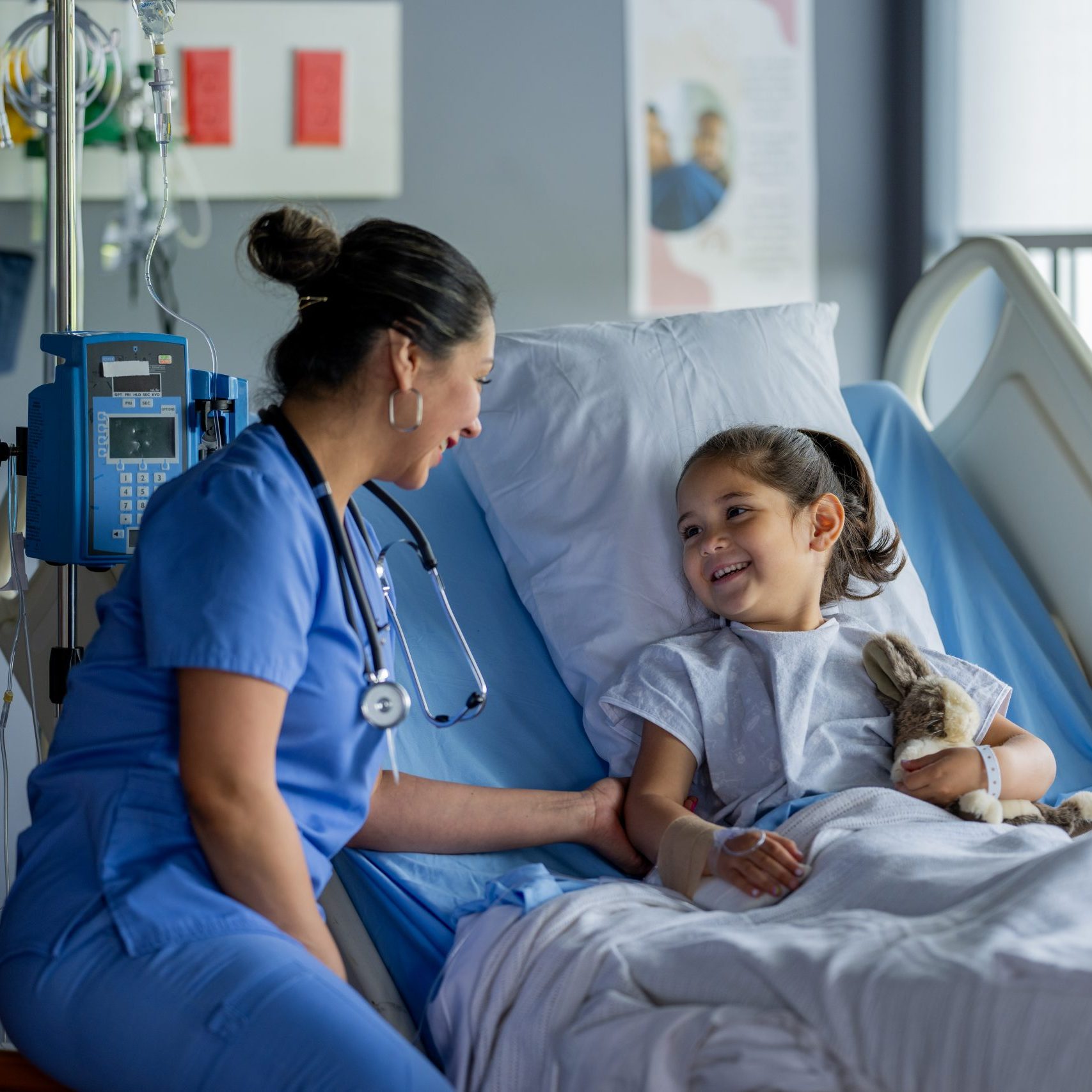 A female nurse of Hispanic decent, makes her rounds to check in on patients after their surgery.  She is sitting with a sweet little girl who is recovering in her hospital bed and cuddling her stuffed animal as she talks with her.