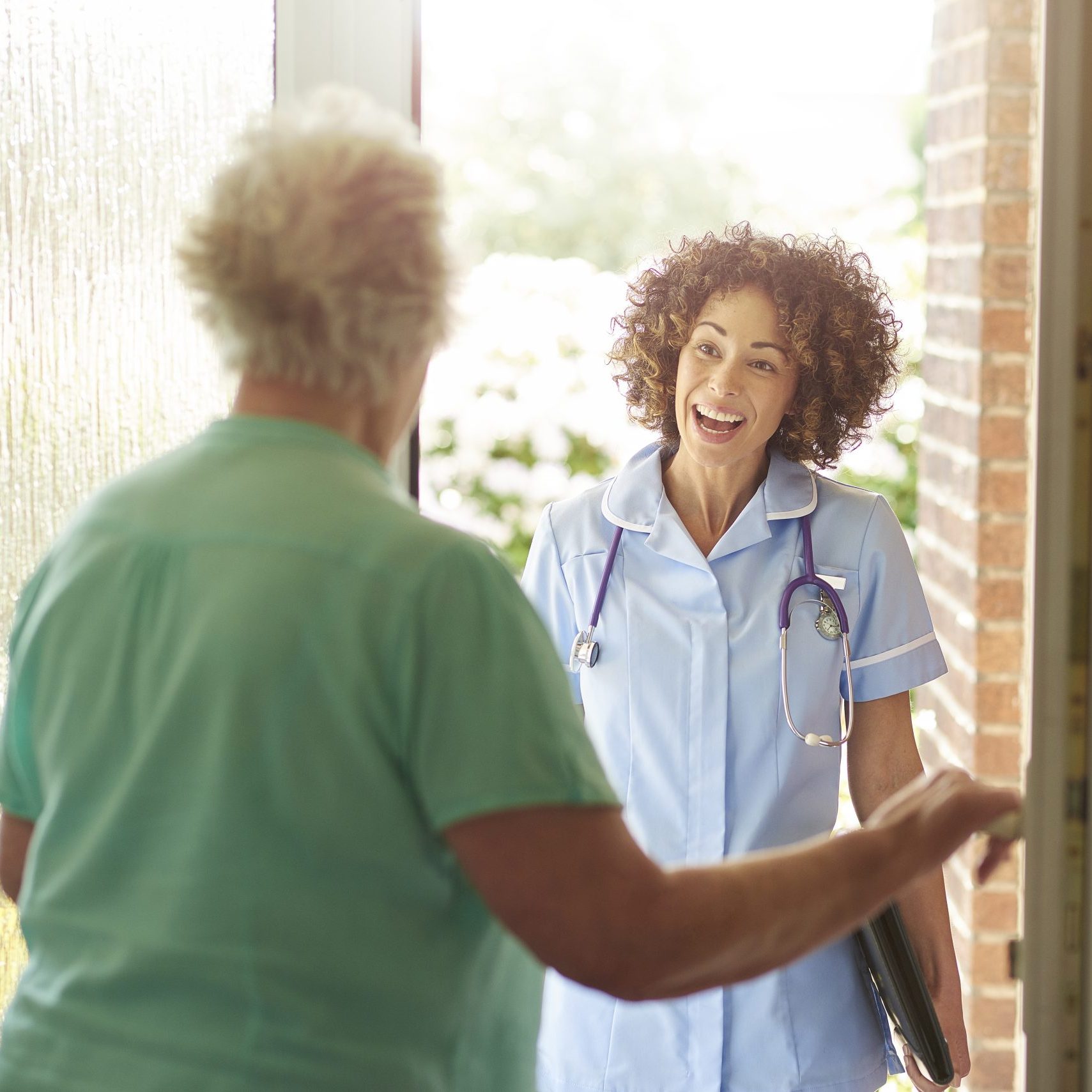 a care worker or medical professional makes a house call to a senior patient at her home . They are standing at the front door as she enters.
