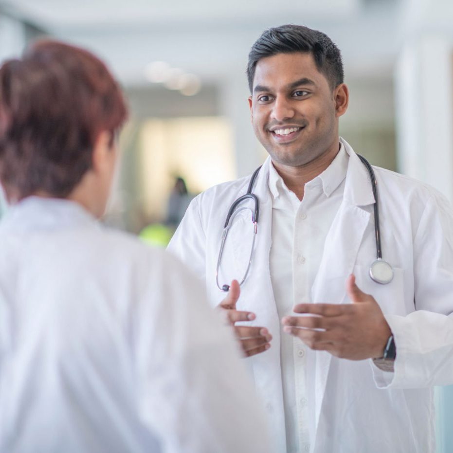 An Indian doctor stands in a hospital hallway discussing his patient's treatment with a female doctor. The man is standing with his hands out in front of him as he uses them to visually help his colleague understand his suggestion.